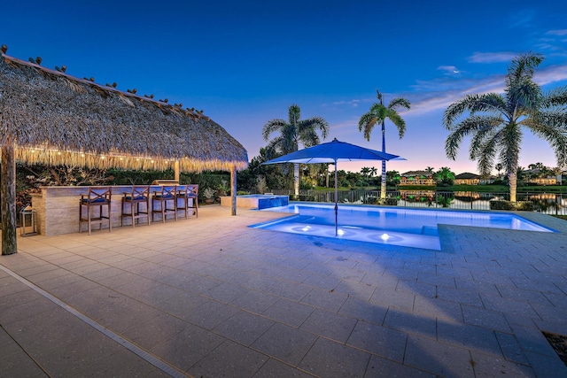 pool at dusk with pool water feature, a water view, an in ground hot tub, a bar, and a patio