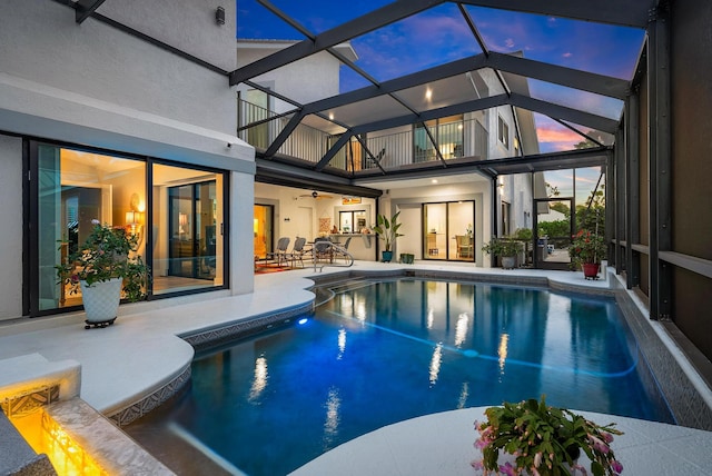 pool at dusk with a patio, glass enclosure, and ceiling fan