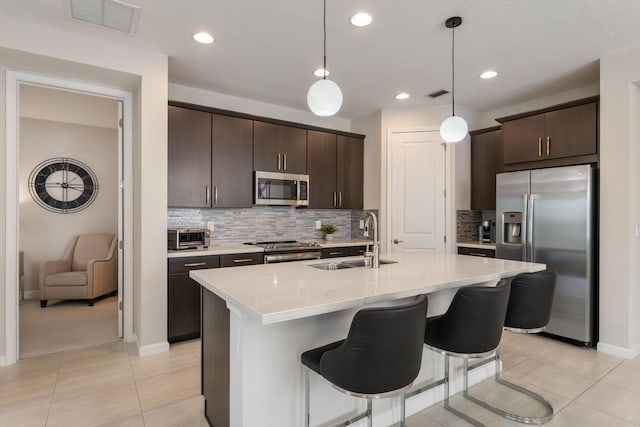 kitchen with sink, appliances with stainless steel finishes, hanging light fixtures, dark brown cabinetry, and an island with sink