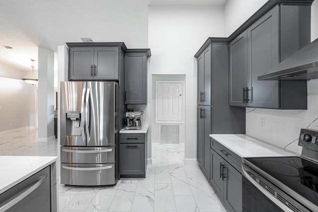 kitchen with gray cabinetry, backsplash, wall chimney exhaust hood, light stone countertops, and stainless steel appliances