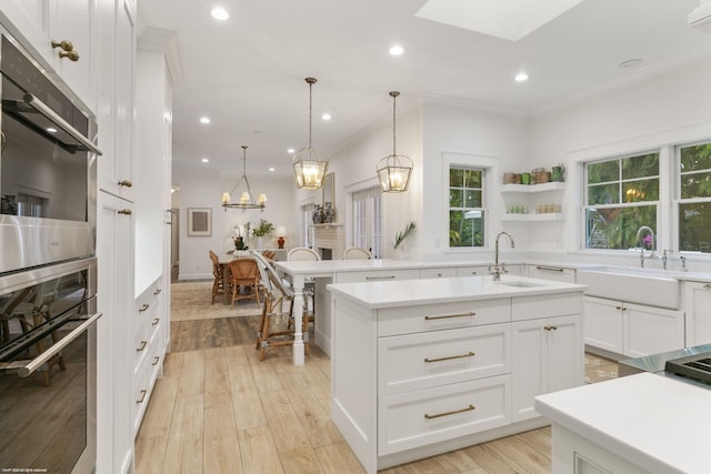 kitchen with double oven, sink, a center island with sink, white cabinetry, and hanging light fixtures