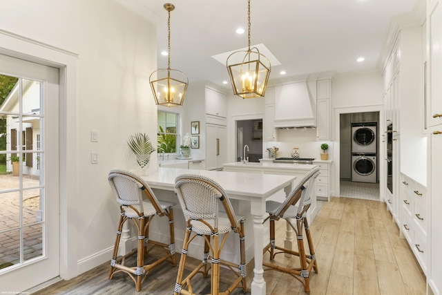 kitchen with white cabinetry, stacked washer / dryer, light hardwood / wood-style floors, pendant lighting, and custom exhaust hood