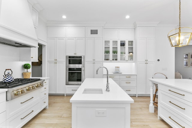 kitchen with sink, stainless steel appliances, premium range hood, an island with sink, and decorative light fixtures