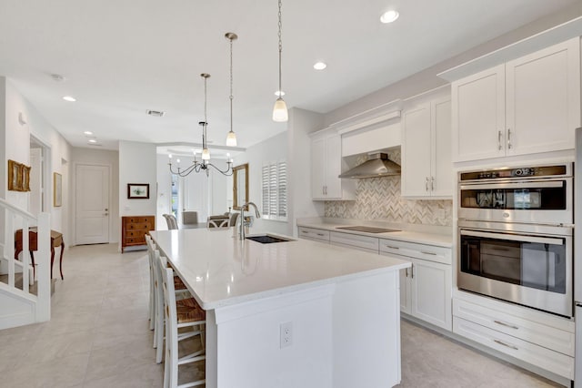 kitchen featuring sink, wall chimney exhaust hood, stainless steel double oven, decorative light fixtures, and a center island with sink