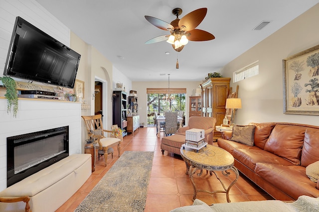 living room featuring light tile patterned floors, a glass covered fireplace, visible vents, and ceiling fan with notable chandelier