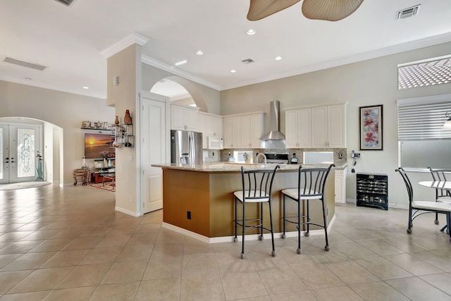 kitchen with wall chimney range hood, stainless steel refrigerator with ice dispenser, an island with sink, light stone counters, and a breakfast bar area