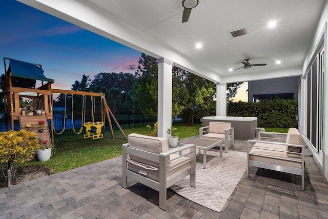 patio terrace at dusk with outdoor lounge area, ceiling fan, a playground, and a hot tub