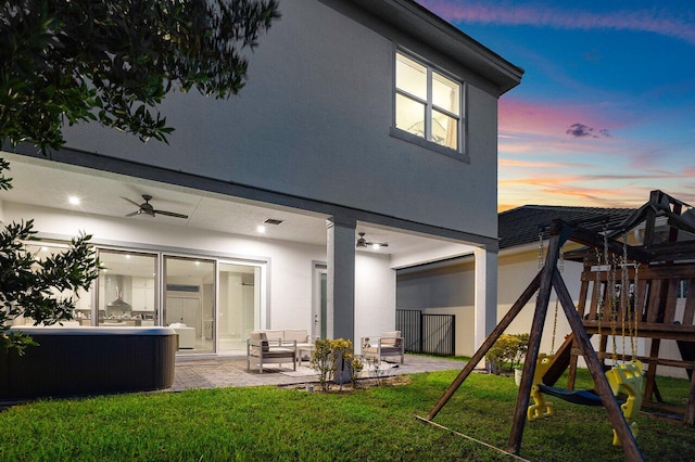 back house at dusk with a playground, ceiling fan, a lawn, and a patio