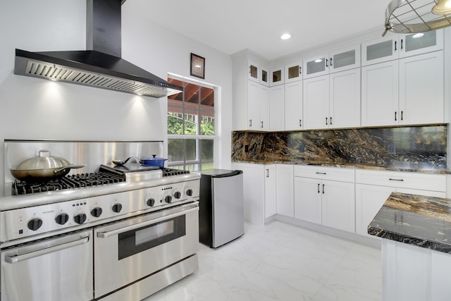 kitchen with high end range, decorative backsplash, wall chimney exhaust hood, dark stone countertops, and white cabinetry