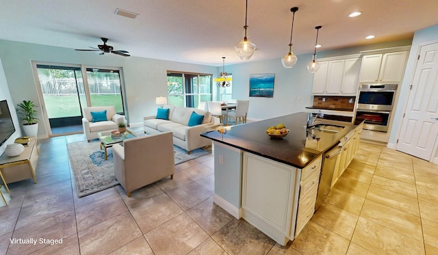 kitchen featuring decorative backsplash, stainless steel double oven, ceiling fan, hanging light fixtures, and an island with sink