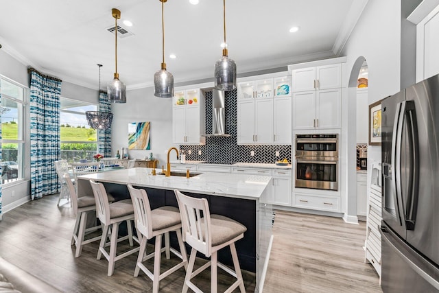 kitchen with wall chimney exhaust hood, stainless steel appliances, sink, white cabinets, and an island with sink