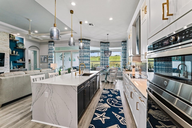 kitchen featuring sink, double oven, pendant lighting, a center island with sink, and white cabinets