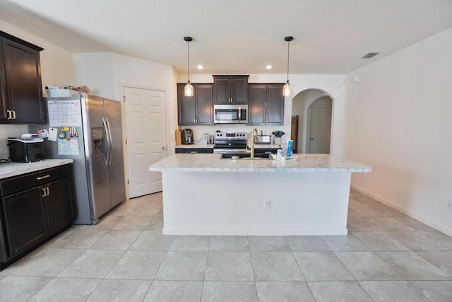 kitchen with light stone counters, dark brown cabinets, stainless steel appliances, a kitchen island with sink, and decorative light fixtures