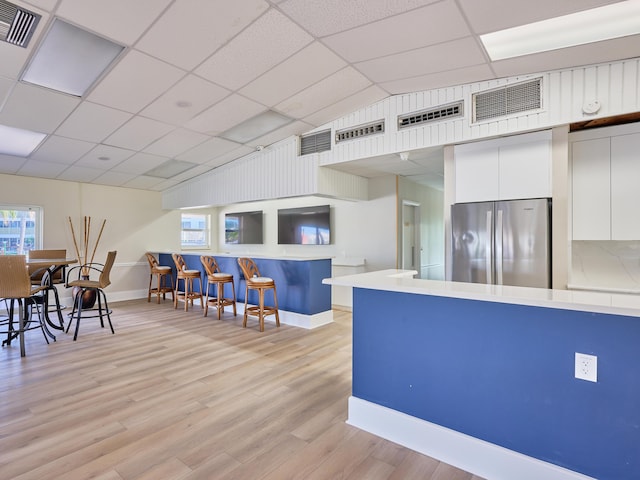 kitchen featuring stainless steel refrigerator, white cabinetry, a drop ceiling, a breakfast bar area, and light wood-type flooring