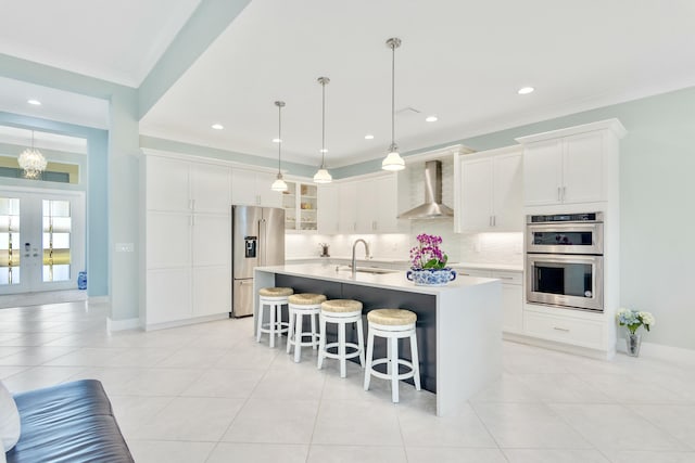 kitchen with white cabinets, wall chimney exhaust hood, an island with sink, decorative light fixtures, and stainless steel appliances