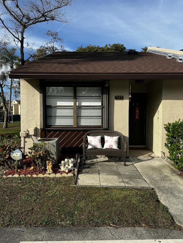 entrance to property featuring a patio area, central AC unit, a shingled roof, and stucco siding