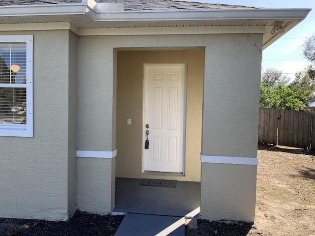 doorway to property featuring stucco siding, fence, and roof with shingles