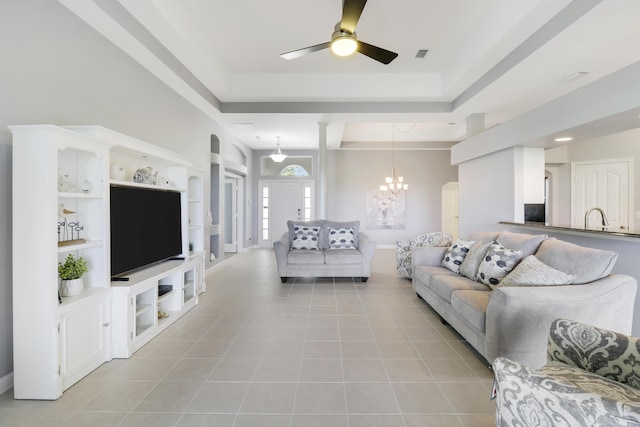 tiled living room with a raised ceiling, built in shelves, sink, and ceiling fan with notable chandelier