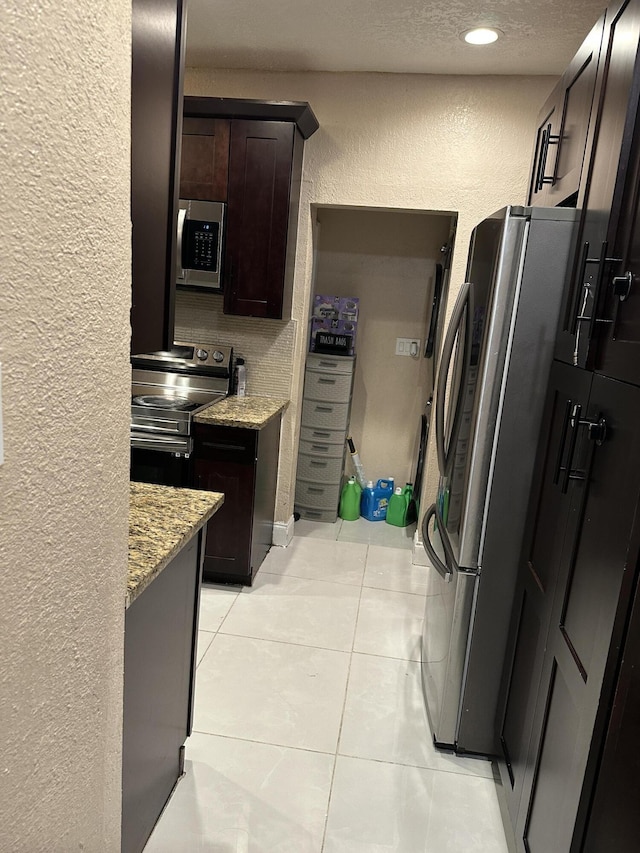 kitchen with light tile patterned floors, a textured ceiling, appliances with stainless steel finishes, light stone counters, and dark brown cabinetry