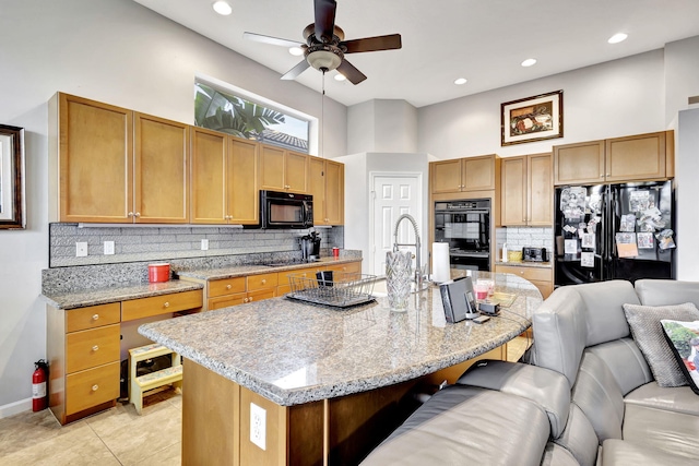 kitchen featuring decorative backsplash, light stone countertops, black appliances, a high ceiling, and an island with sink