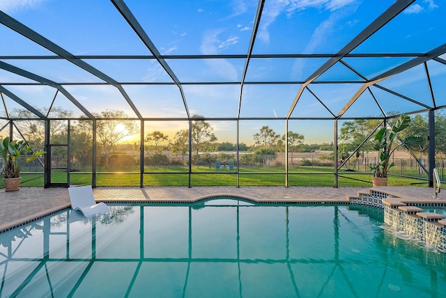 pool at dusk with glass enclosure, a jacuzzi, a patio, a yard, and pool water feature