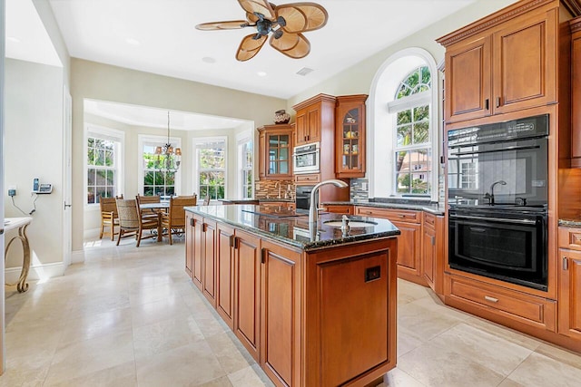 kitchen featuring a center island with sink, ceiling fan with notable chandelier, decorative backsplash, dark stone countertops, and double oven