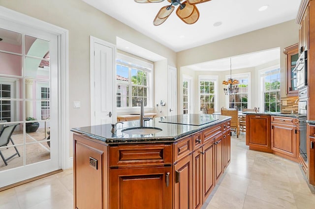 kitchen featuring a kitchen island with sink, black electric stovetop, sink, hanging light fixtures, and dark stone countertops