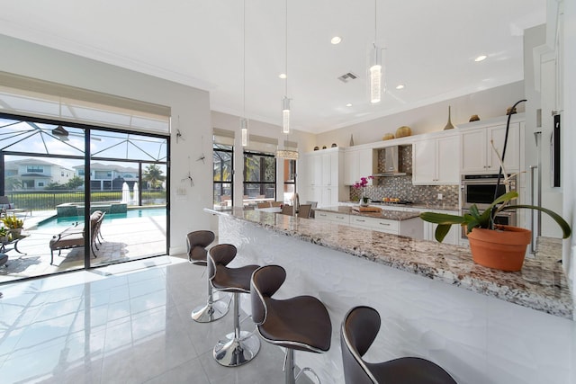 kitchen featuring light stone countertops, wall chimney exhaust hood, tasteful backsplash, decorative light fixtures, and white cabinets