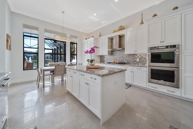 kitchen with pendant lighting, wall chimney range hood, light stone counters, white cabinetry, and stainless steel appliances