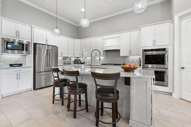 kitchen with custom range hood, hanging light fixtures, appliances with stainless steel finishes, white cabinets, and an island with sink