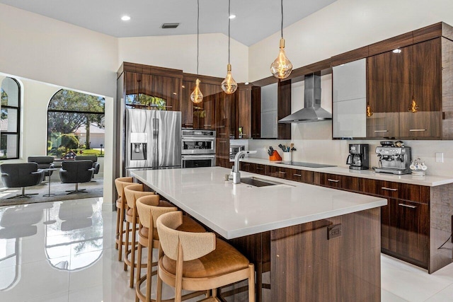 kitchen with appliances with stainless steel finishes, a kitchen island with sink, sink, wall chimney range hood, and hanging light fixtures