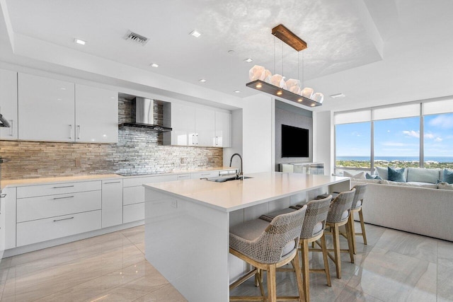 kitchen with wall chimney exhaust hood, a kitchen island with sink, sink, pendant lighting, and white cabinets