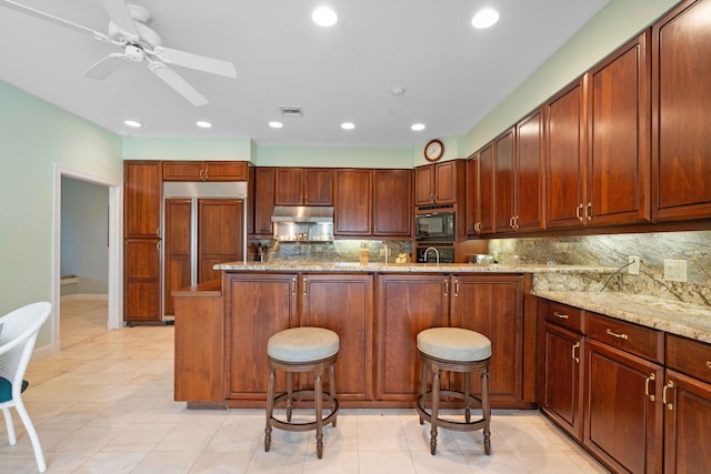 kitchen with a breakfast bar, backsplash, built in appliances, ceiling fan, and light stone countertops