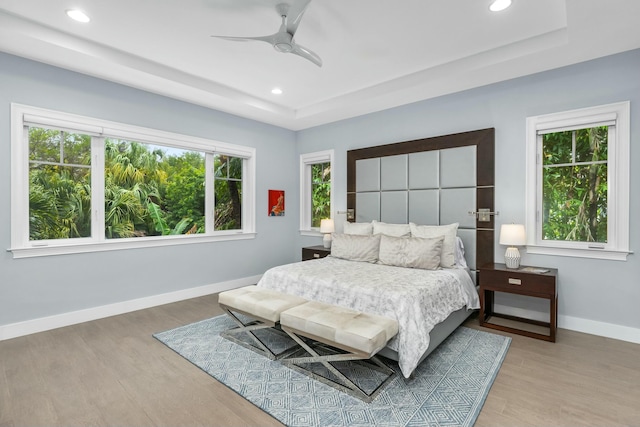 bedroom with ceiling fan, a tray ceiling, light hardwood / wood-style floors, and multiple windows