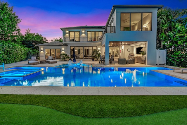 back house at dusk featuring a pool with hot tub, an outdoor kitchen, a patio area, and a balcony