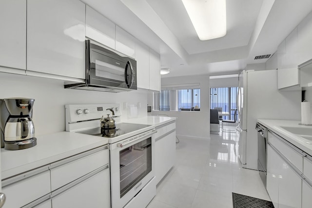 kitchen with a raised ceiling, white electric range, stainless steel dishwasher, light tile patterned flooring, and white cabinetry