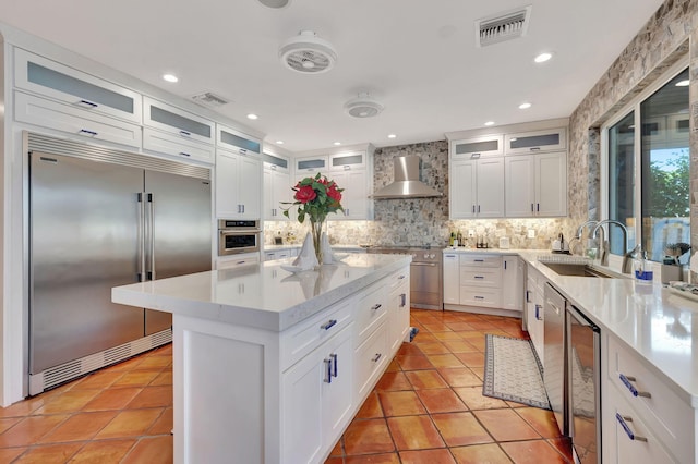 kitchen featuring wall chimney exhaust hood, a kitchen island, backsplash, white cabinetry, and appliances with stainless steel finishes