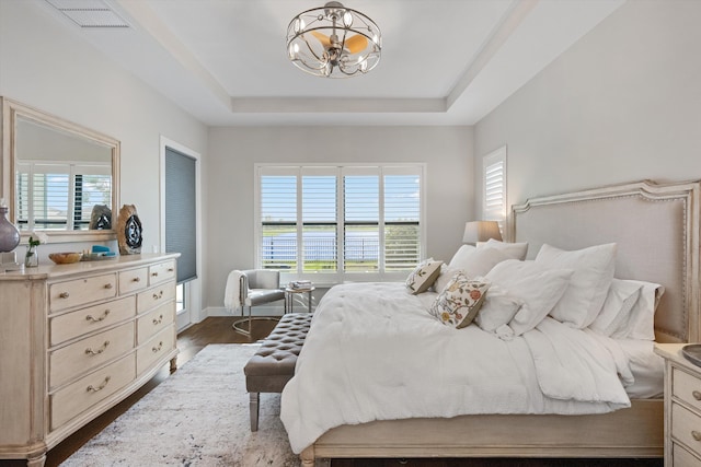 bedroom featuring dark hardwood / wood-style flooring, a water view, a raised ceiling, and a notable chandelier