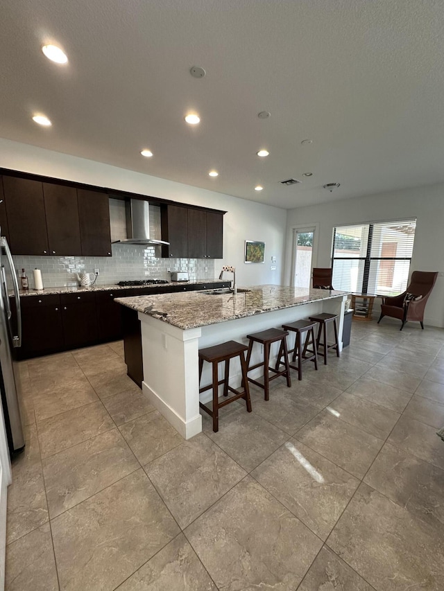 kitchen featuring a kitchen breakfast bar, wall chimney range hood, an island with sink, light stone counters, and stainless steel refrigerator