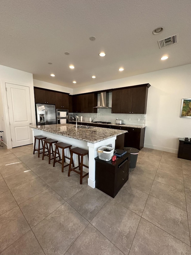 kitchen featuring a kitchen island with sink, wall chimney range hood, dark brown cabinetry, light stone counters, and stainless steel fridge with ice dispenser