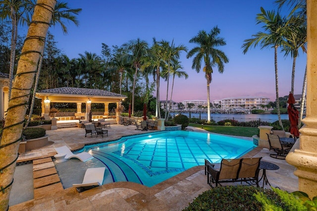 pool at dusk with a gazebo, a patio, a water view, and an outdoor living space