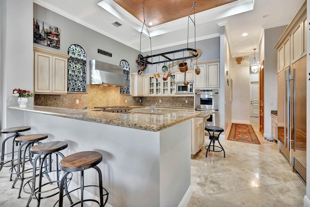 kitchen featuring stainless steel appliances, wall chimney range hood, a kitchen breakfast bar, cream cabinets, and a tray ceiling