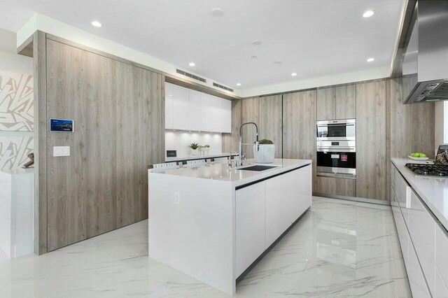 kitchen with double wall oven, wall chimney exhaust hood, sink, a center island with sink, and white cabinets