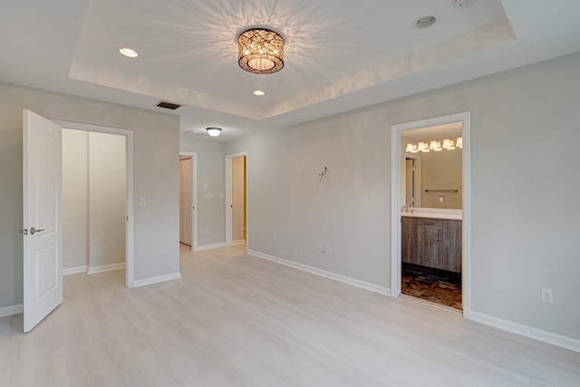 unfurnished bedroom with a raised ceiling, ensuite bathroom, and light hardwood / wood-style floors