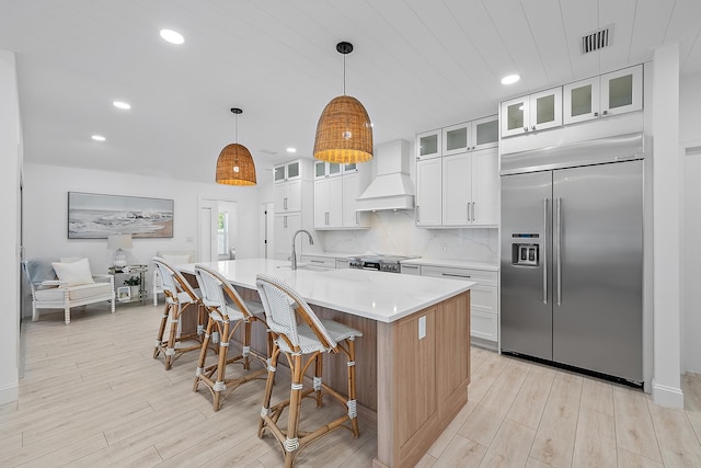 kitchen with custom exhaust hood, stainless steel built in refrigerator, sink, decorative light fixtures, and an island with sink