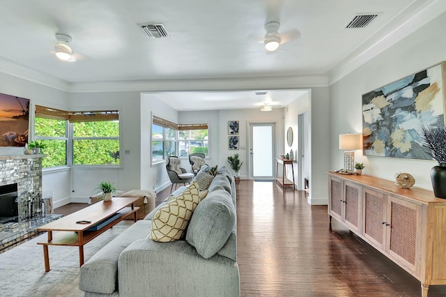 living room featuring a stone fireplace, ceiling fan, crown molding, and dark wood-type flooring