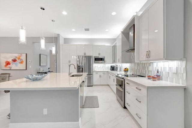 kitchen featuring wall chimney range hood, decorative light fixtures, a kitchen island with sink, white cabinets, and appliances with stainless steel finishes