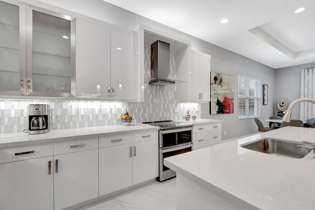 kitchen featuring double oven range, backsplash, wall chimney range hood, sink, and white cabinetry