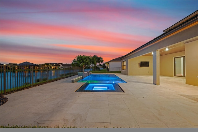 pool at dusk featuring a patio area, a water view, and an in ground hot tub