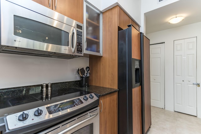 kitchen with glass insert cabinets, dark stone counters, light tile patterned floors, brown cabinets, and appliances with stainless steel finishes